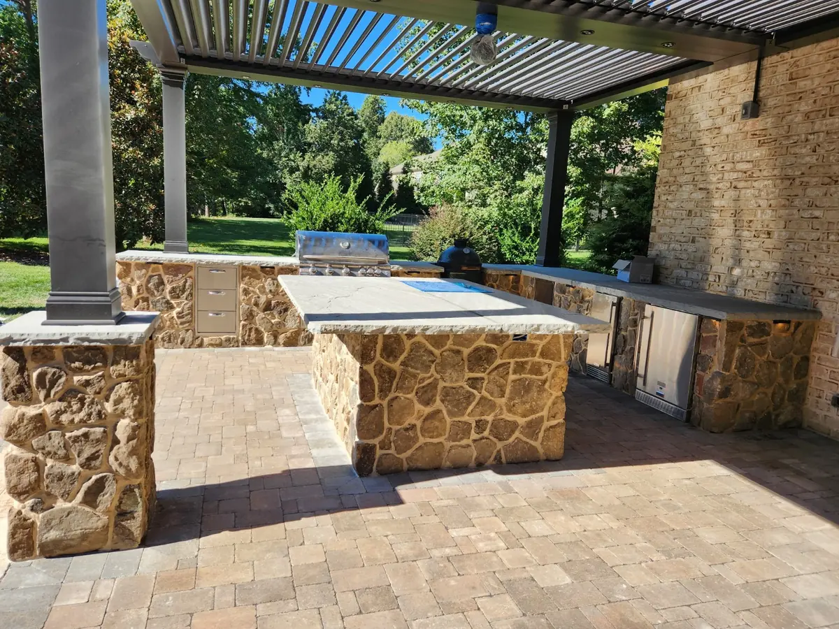 U-shaped stone outdoor kitchen with built-in grill, refrigerator, and charcoal grill under a pergola